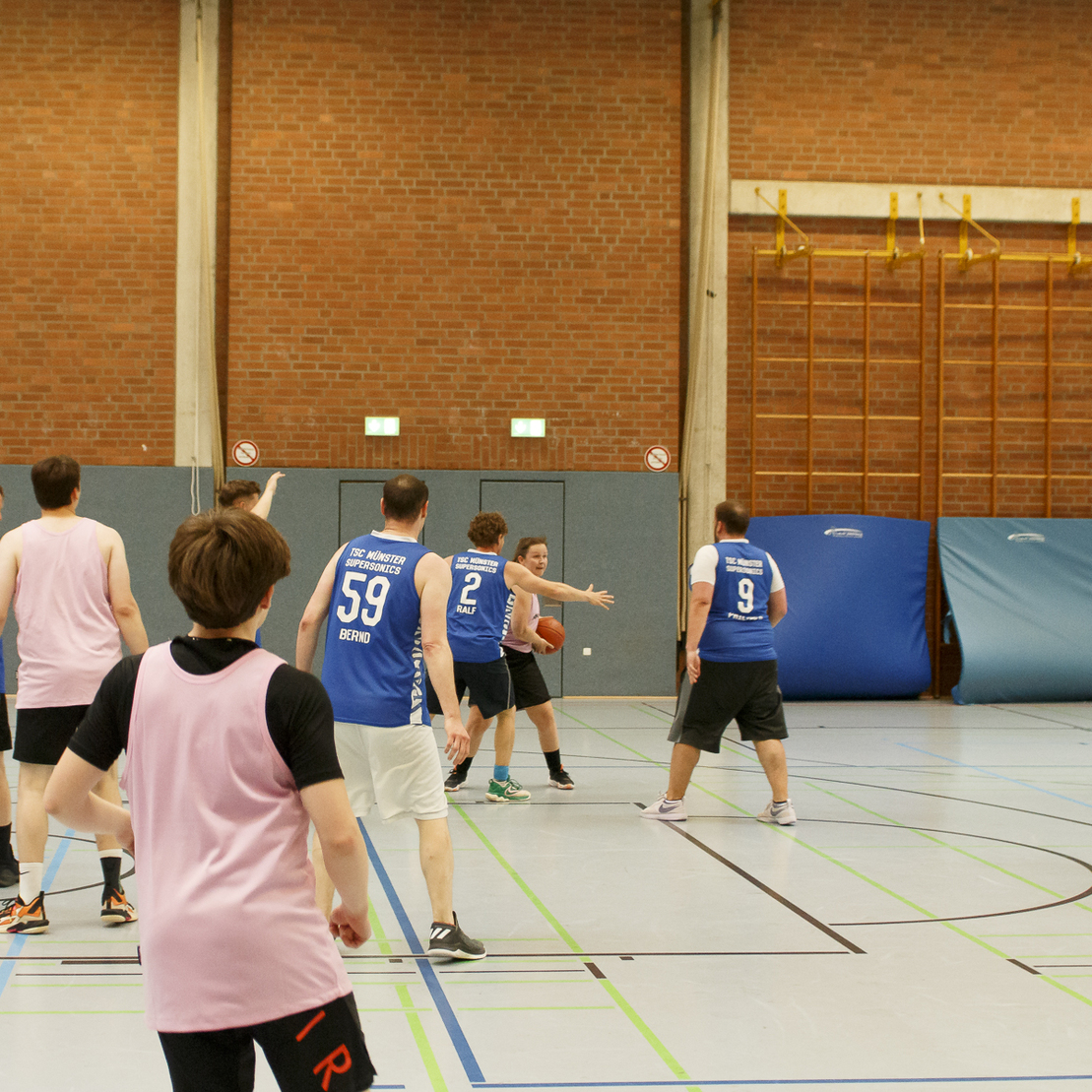 Basketballspiel in einer Halle mit Spielern in blauen und pinken Trikots, die um den Ball k&auml;mpfen.
