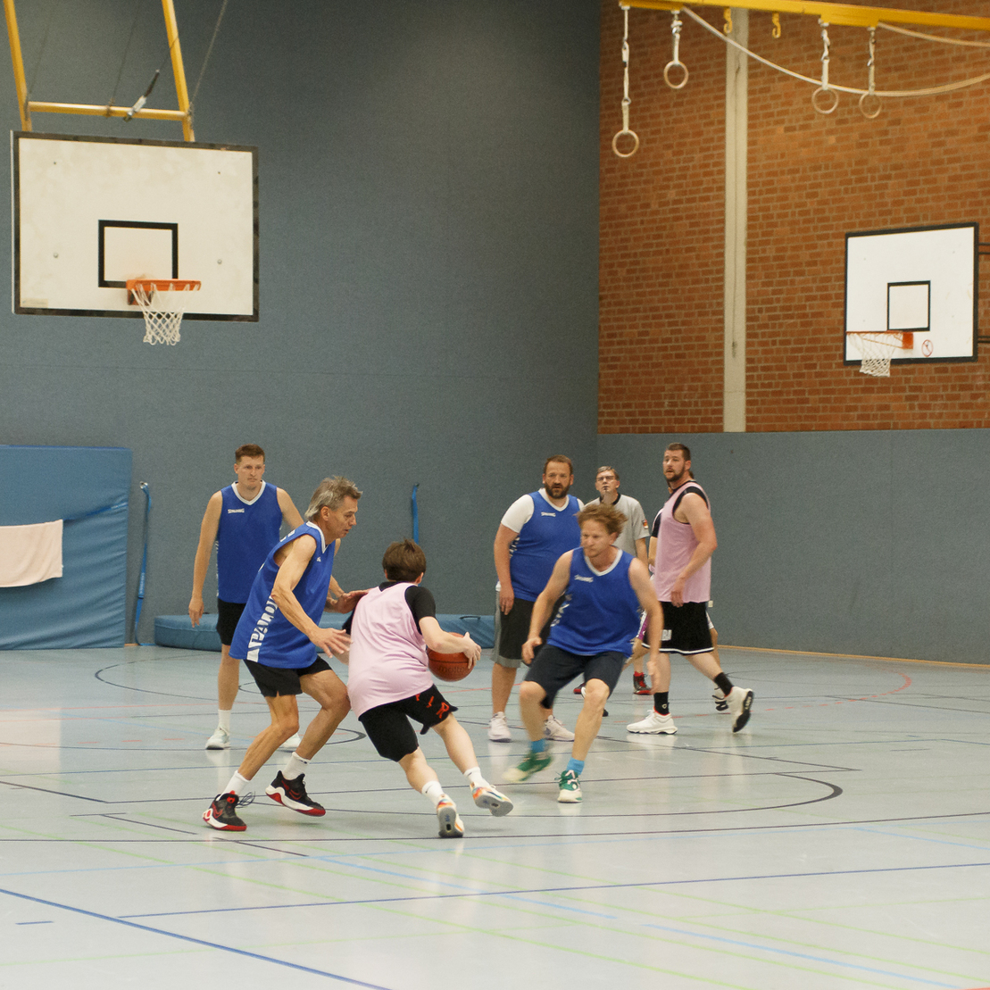 Zehn M&auml;nner spielen Basketball in einer Sporthalle, einige tragen Trikots in Blau und Pink.