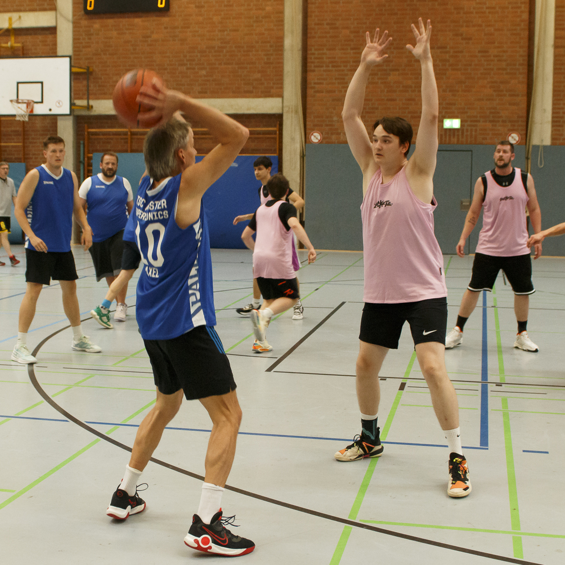 Mehrere M&auml;nner spielen Basketball in einer Sporthalle; ein Spieler wirft den Ball, w&auml;hrend ein anderer ihn verteidigt.