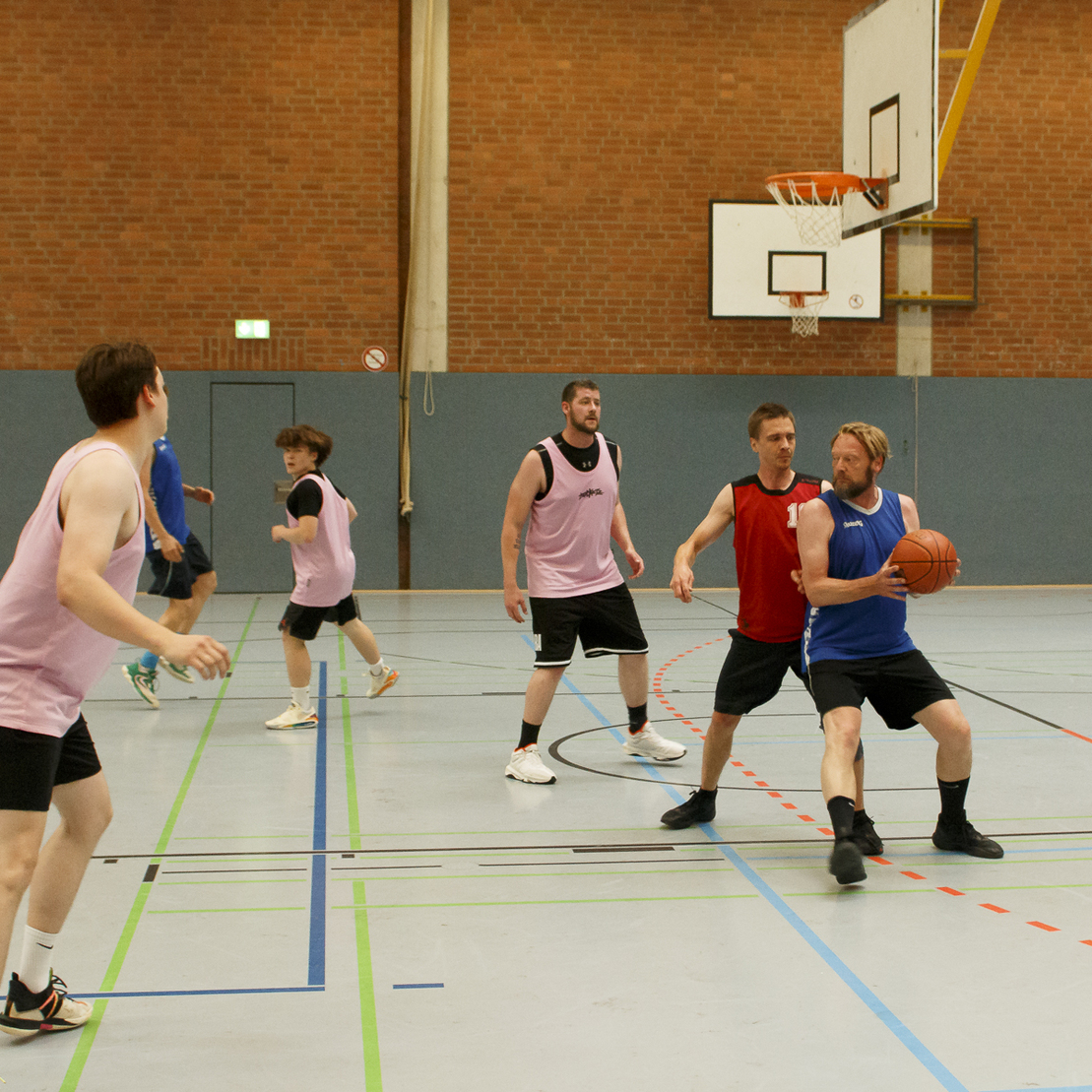 F&uuml;nf Basketballspieler in T-Shirts stehen auf dem Spielfeld, einer dribbelt den Ball vor einem Verteidiger.