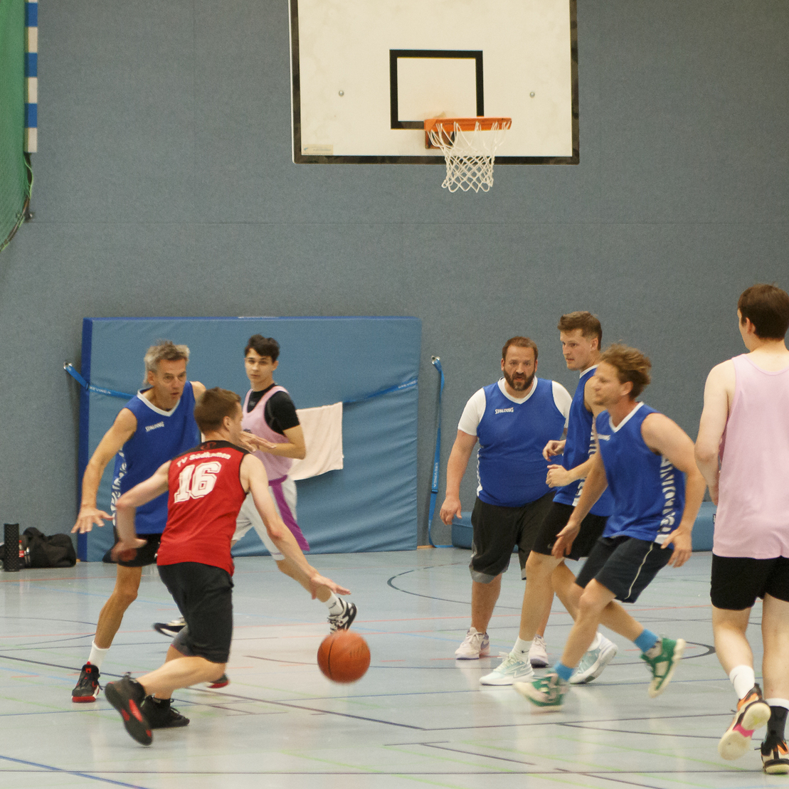 Spielszene im Basketball mit sechs Spielern in blauen und roten Trikots auf dem Court unter einem Basketballkorb.