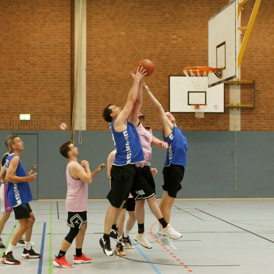 Basketballspiel in einer Halle: Zwei Spieler springen f&uuml;r den Ball &uuml;ber anderen Spielern in rosa Trikots.
