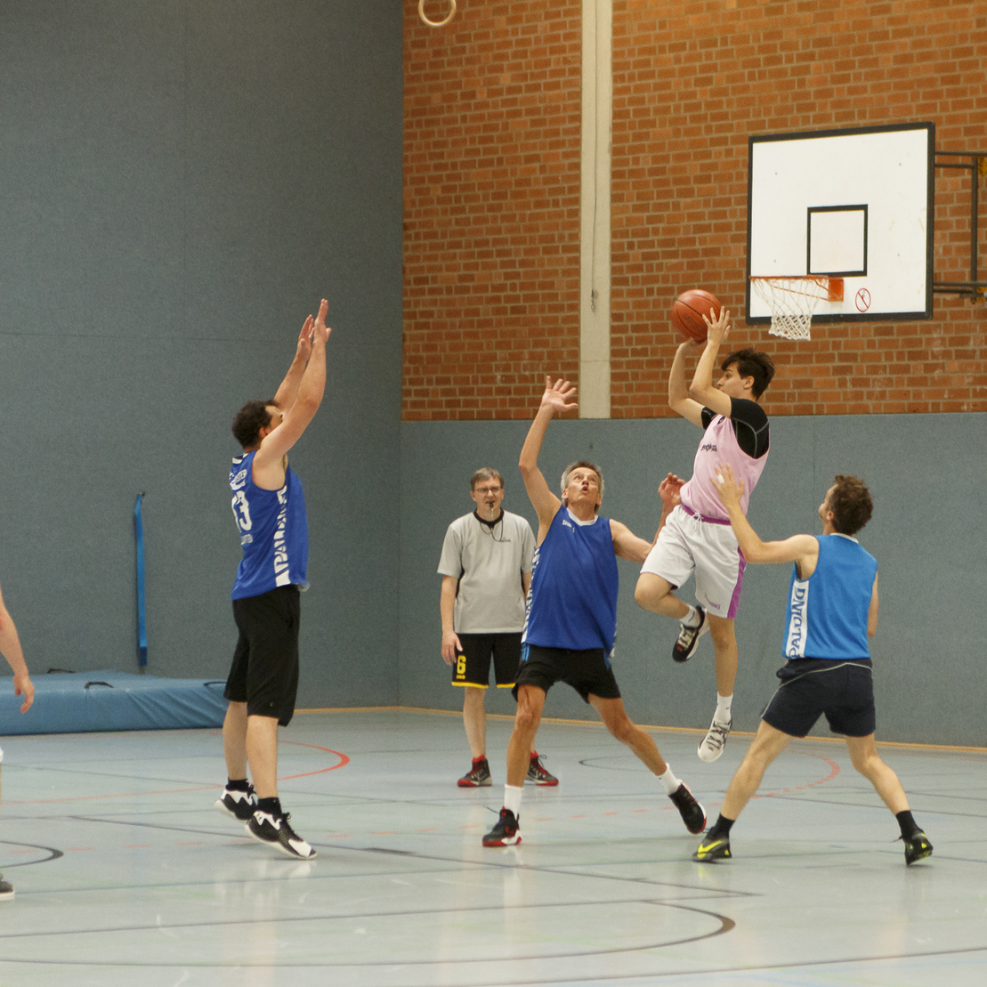 Spieler in blauen und schwarzen Trikots greifen in einer Sporthalle nach dem Basketball w&auml;hrend eines Spiels.