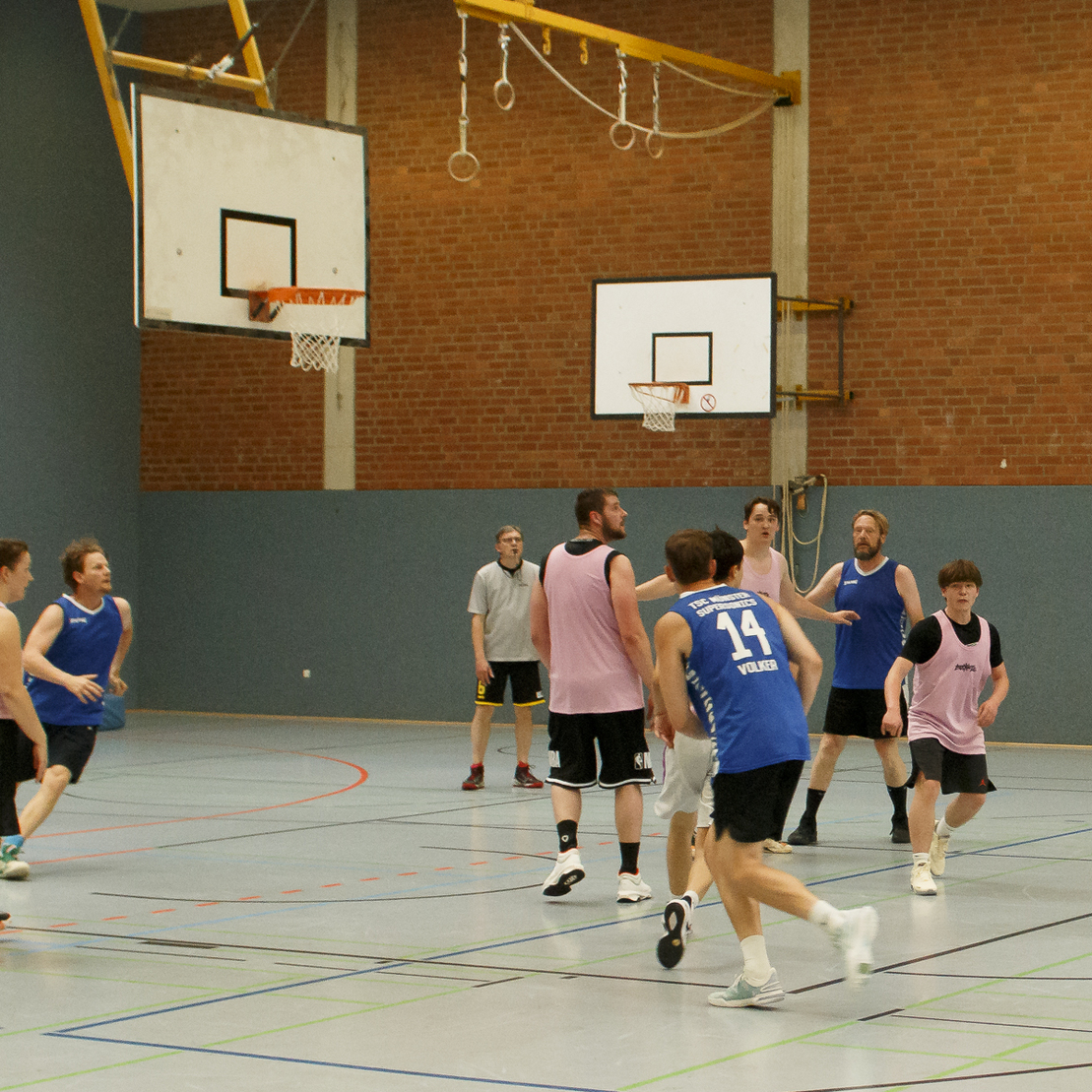 Verschiedene Personen spielen Basketball in einer Sporthalle, ein Spieler wirft den Ball auf den Korb.