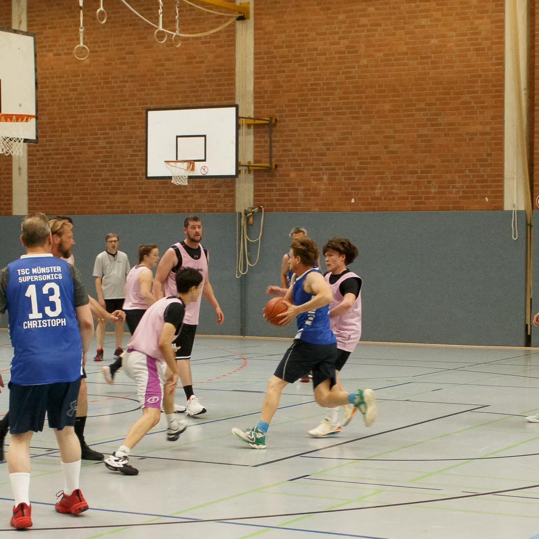 Szenenbild eines Basketballspiels in einer Sporthalle, mit Spielern in Trikots und einem Ball in der Mitte des Geschehens.