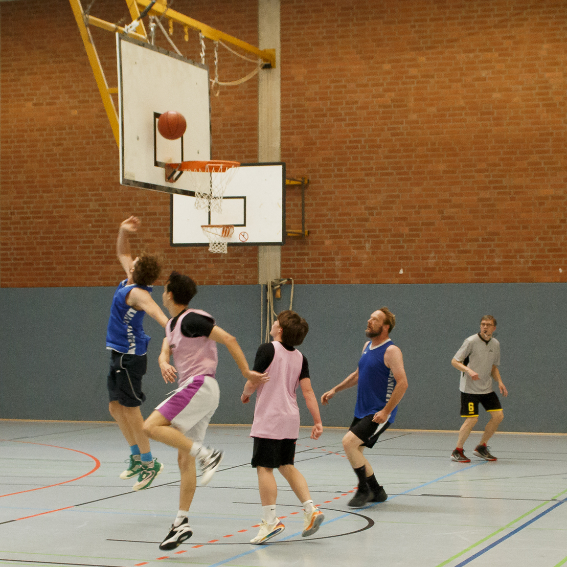 Spielende Basketballspieler im Sprung, w&auml;hrend ein Ball auf den Korb zielt, in einer Sporthalle mit Holzlattenwand.
