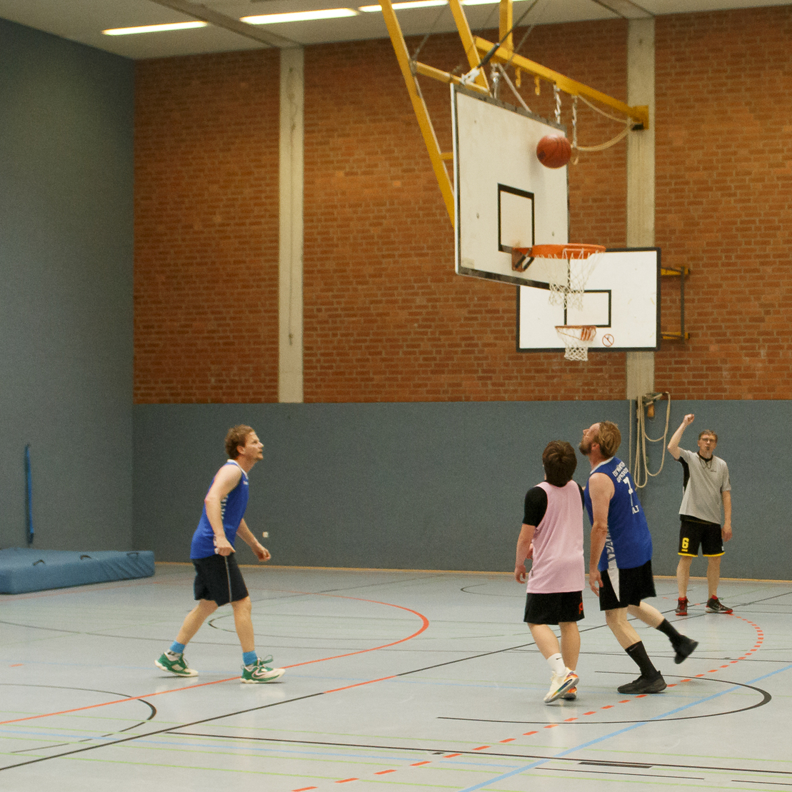 F&uuml;nf Personen spielen Basketball in einer Halle, ein Spieler wirft den Ball Richtung Korb.