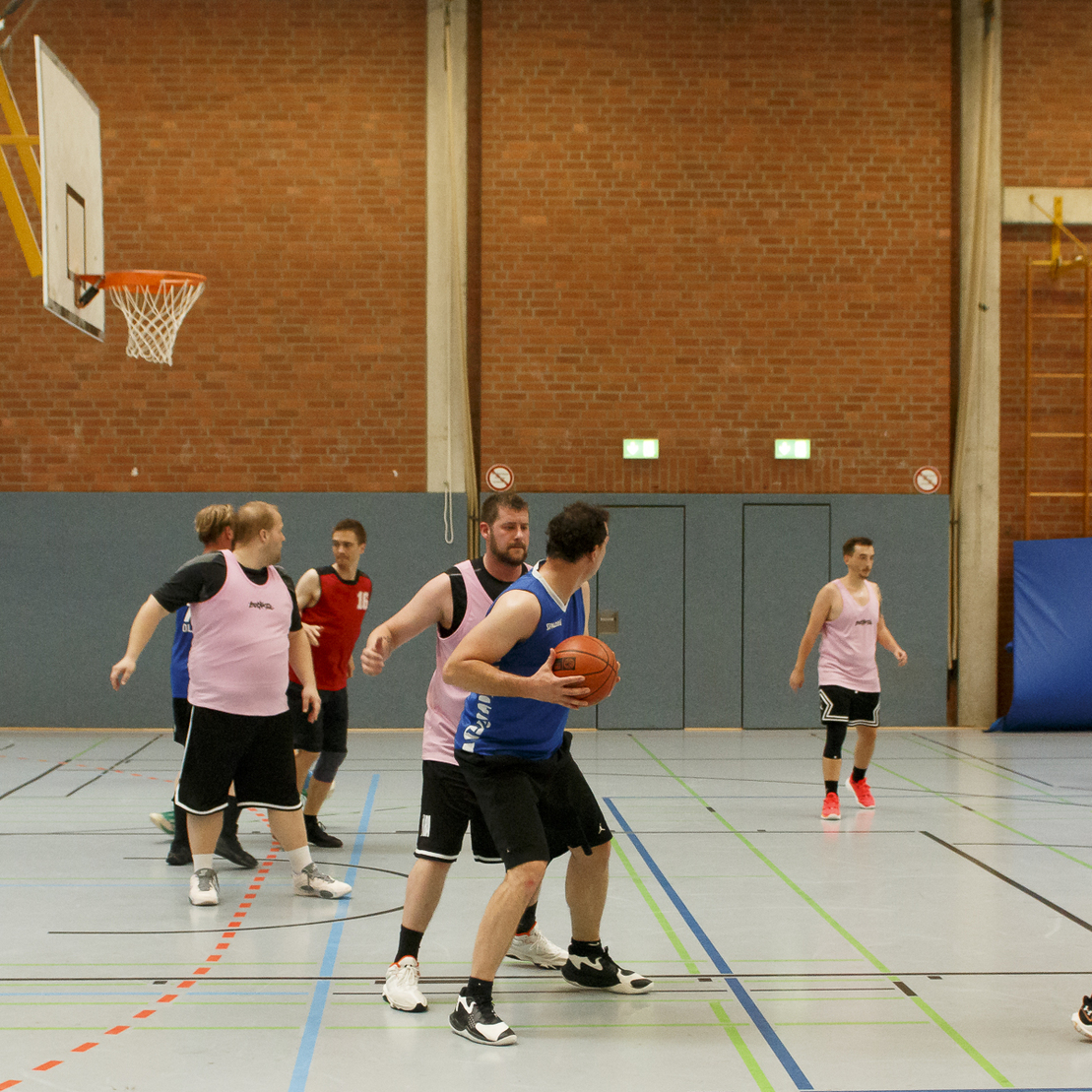 Zwei Mannschaften spielen Basketball in einer Halle; ein Spieler mit Ball wird von einem anderen angegriffen.