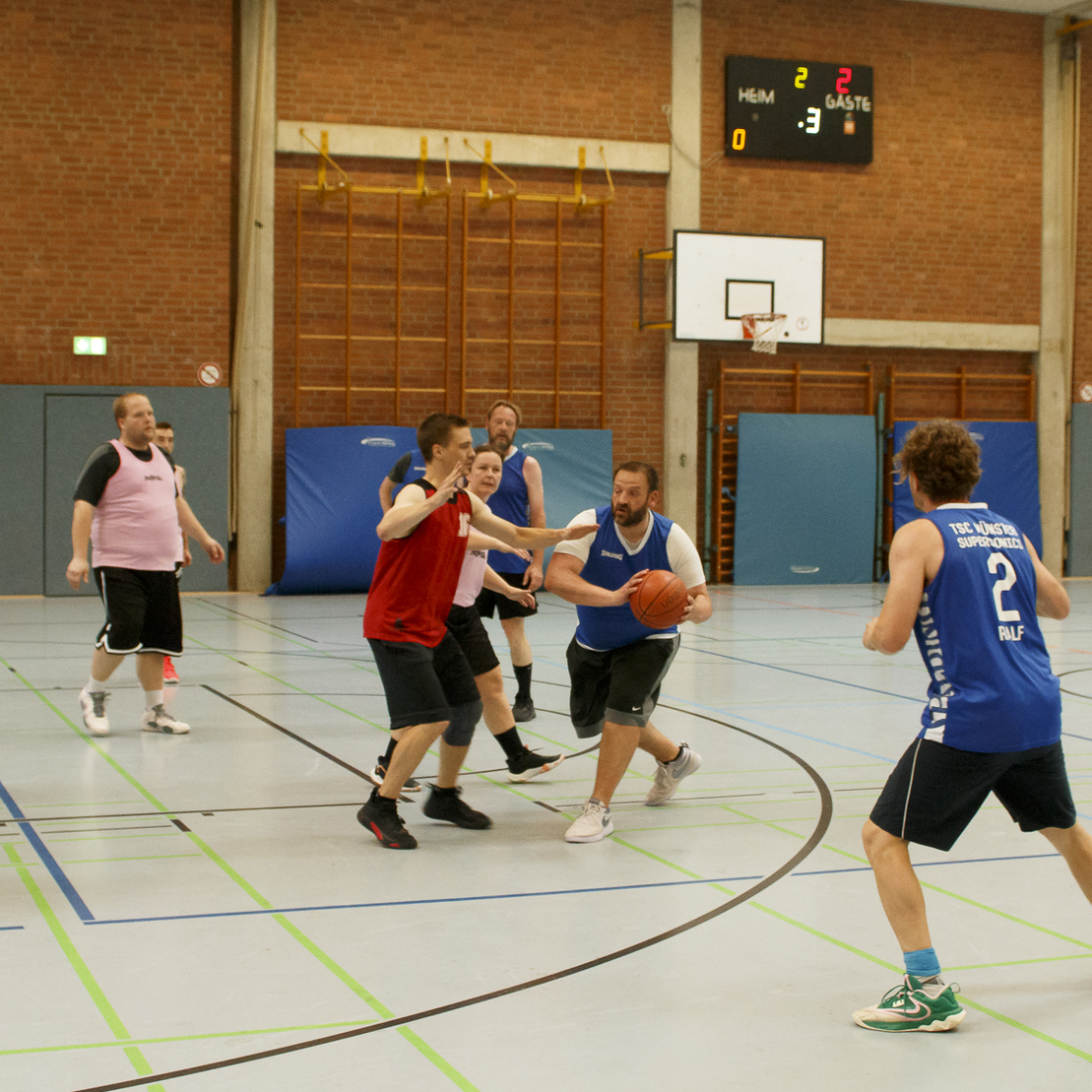 Basketballspiel in einer Sporthalle mit Spielern in Trikots, die um den Ball k&auml;mpfen. Punkte stehen 2 zu 3.