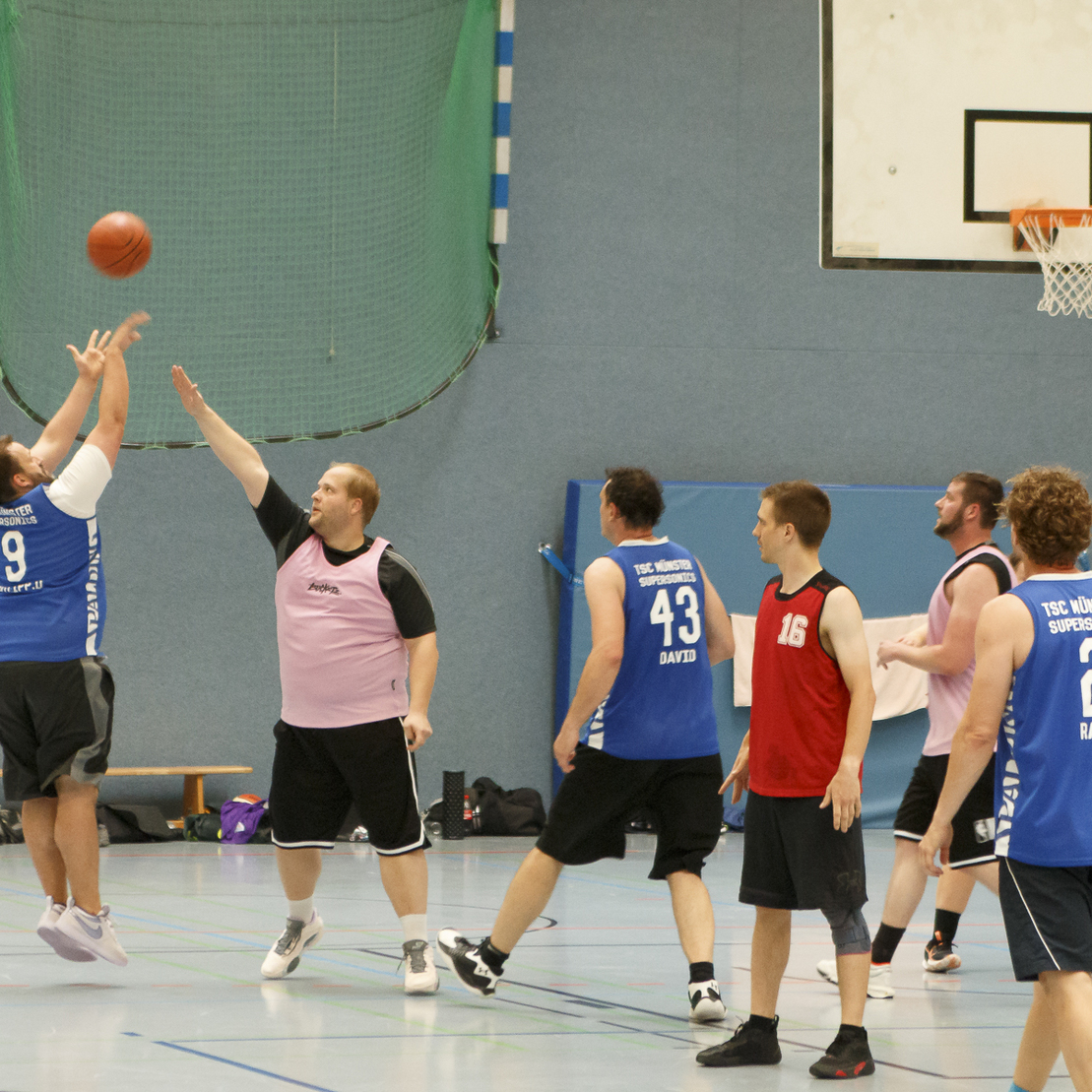 Spielszene in einer Halle: Ein Spieler wirft den Basketball zum Korb, w&auml;hrend ein Gegenspieler versucht, ihn zu blocken.
