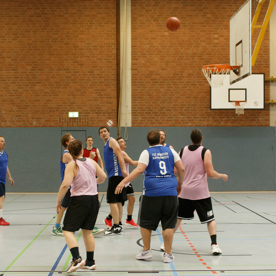 Basketballspieler in Sportkleidung werfen Ball Richtung Korb in einer Halle mit Holz- und Klinkerboden.