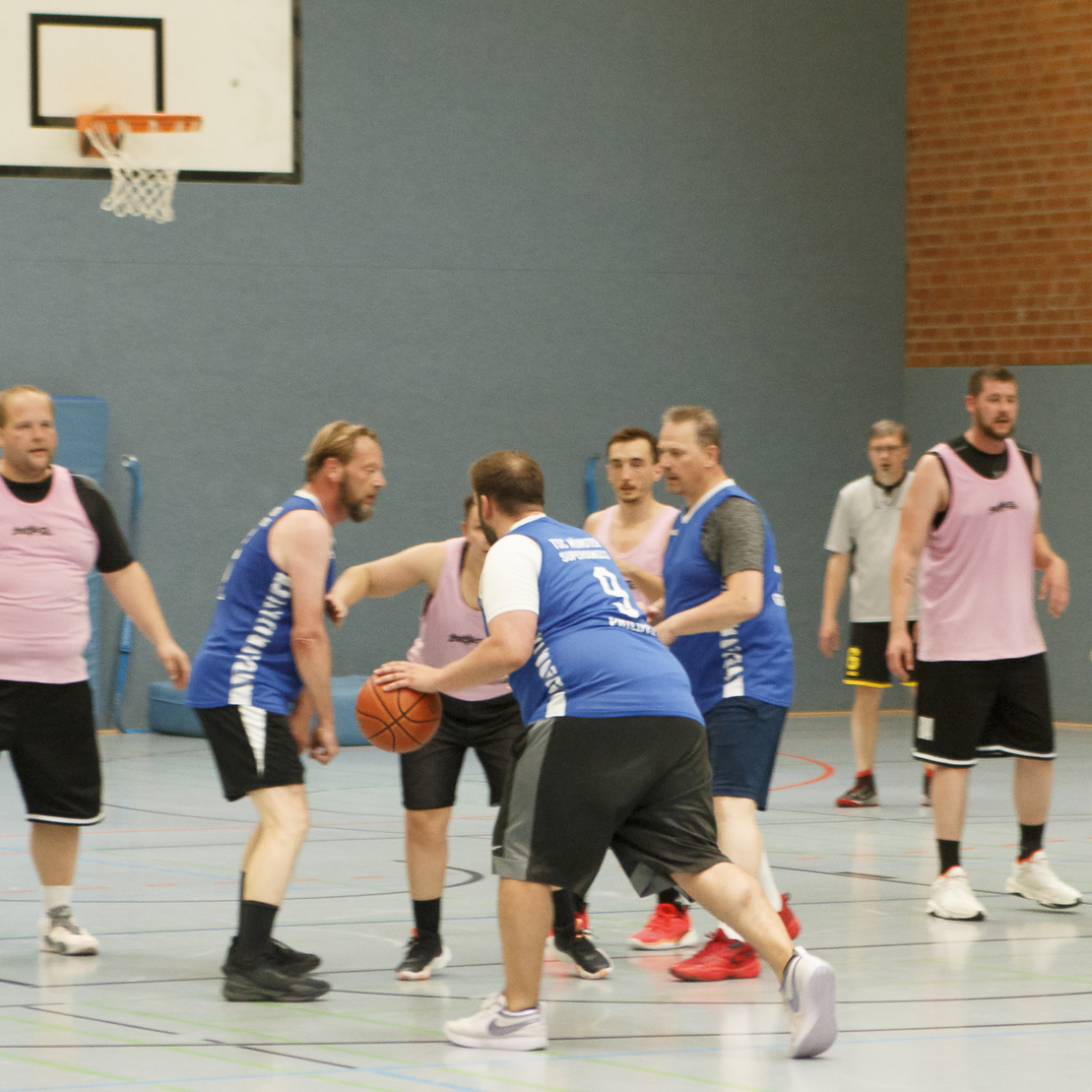 Basketballspieler in Trikots k&auml;mpfen um den Ball auf dem Pad. Der Korb ist im Hintergrund sichtbar.