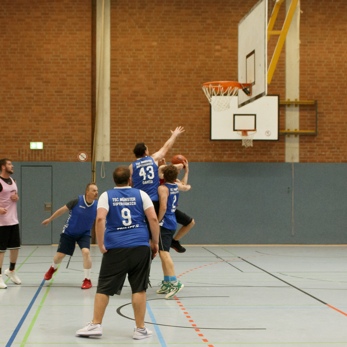 Basketballspieler springen in einer Halle, um den Ball an den Korb zu werfen, w&auml;hrend andere zuschauen.