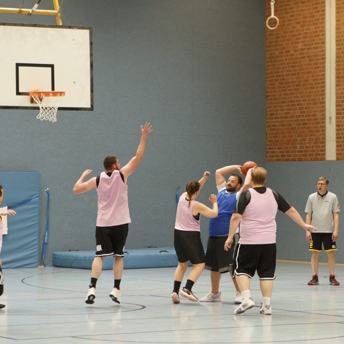 Zwei Basketballmannschaften spielen in einer Halle; ein Spieler wirft den Ball auf den Korb.