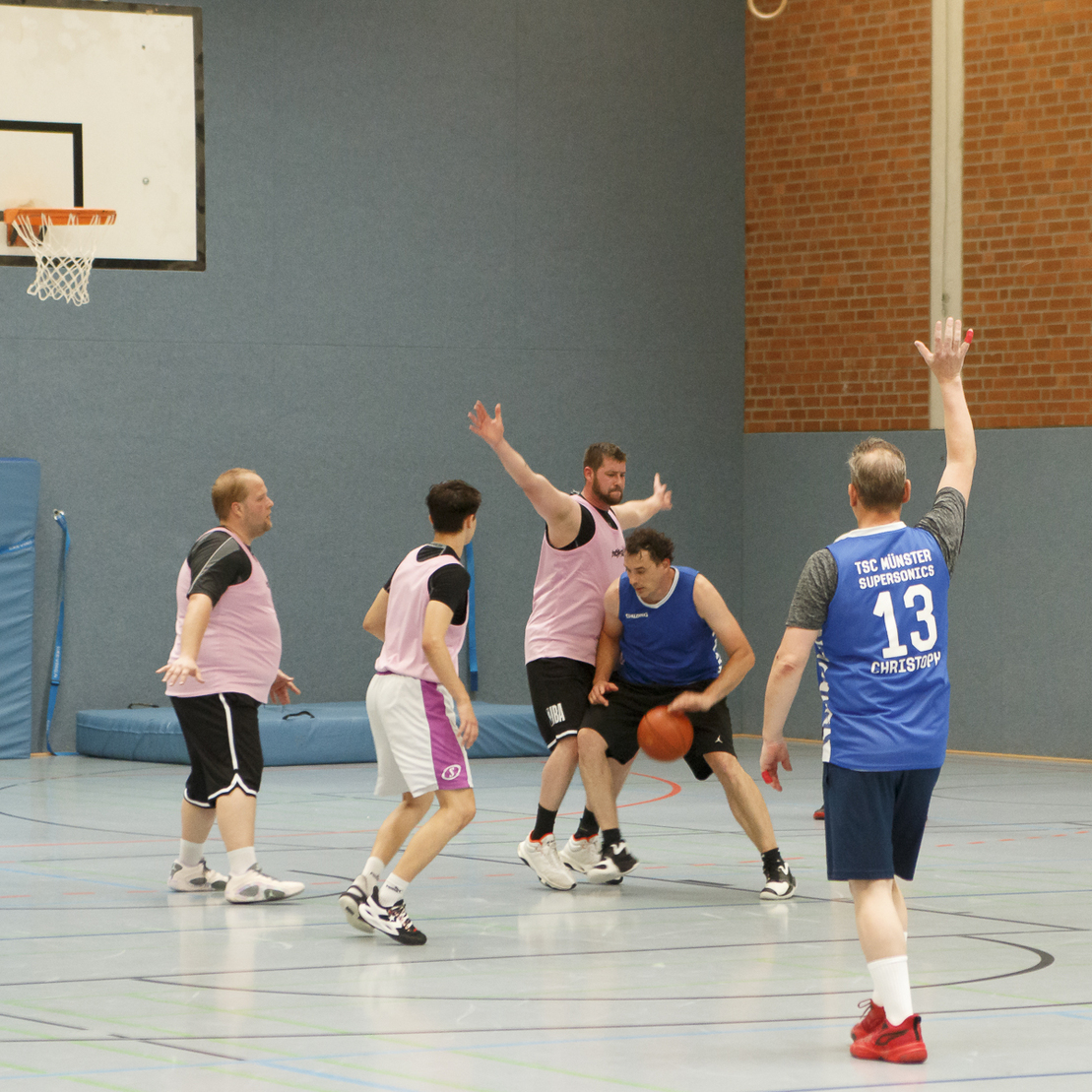 Sieben M&auml;nner in Sportkleidung spielen Basketball in einer Halle, ein Spieler dribbelt den Ball.