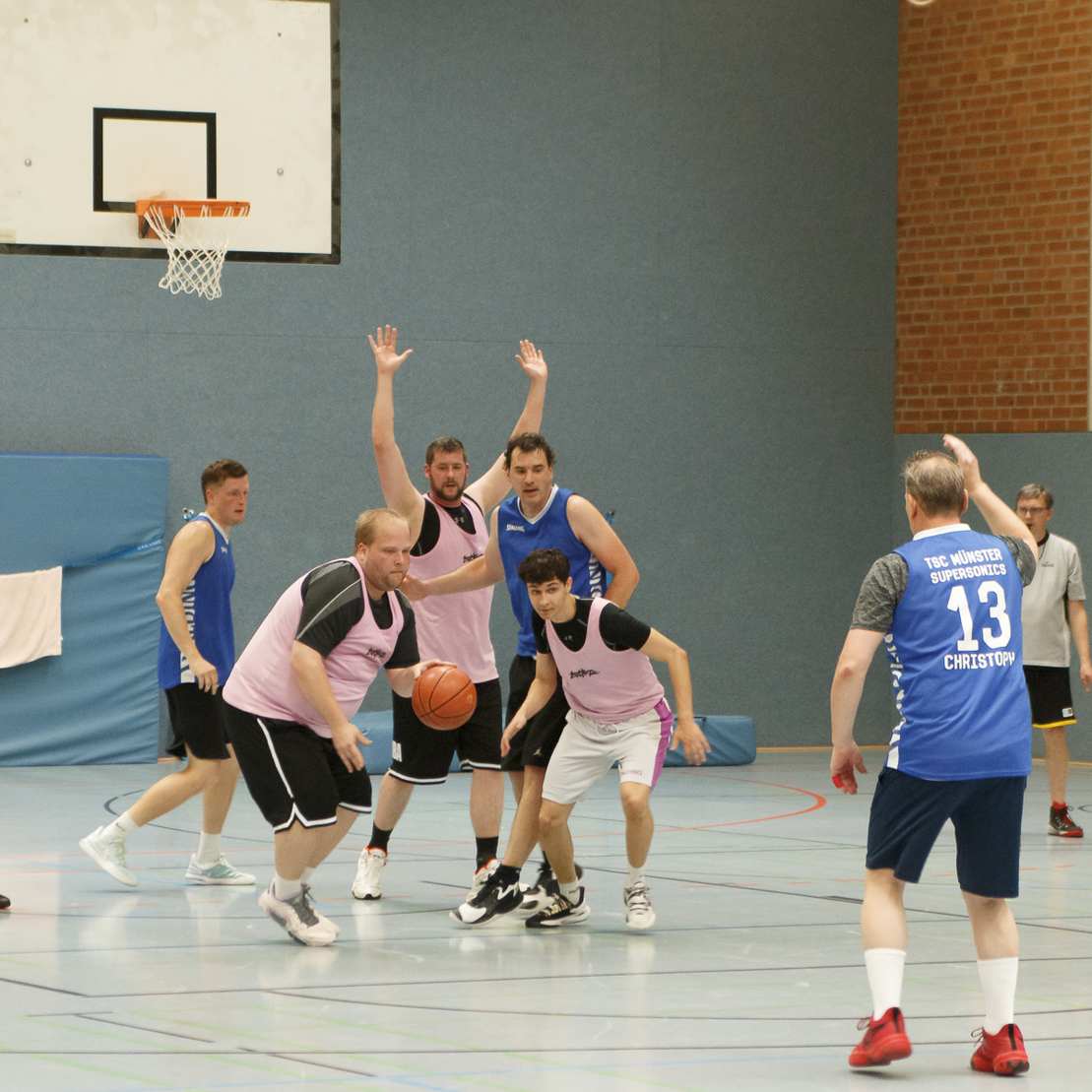 Basketballspiel in einer Halle mit Spielern in bunten Trikots, einer dribbelt den Ball, andere bereiten sich auf den Wurf vor.