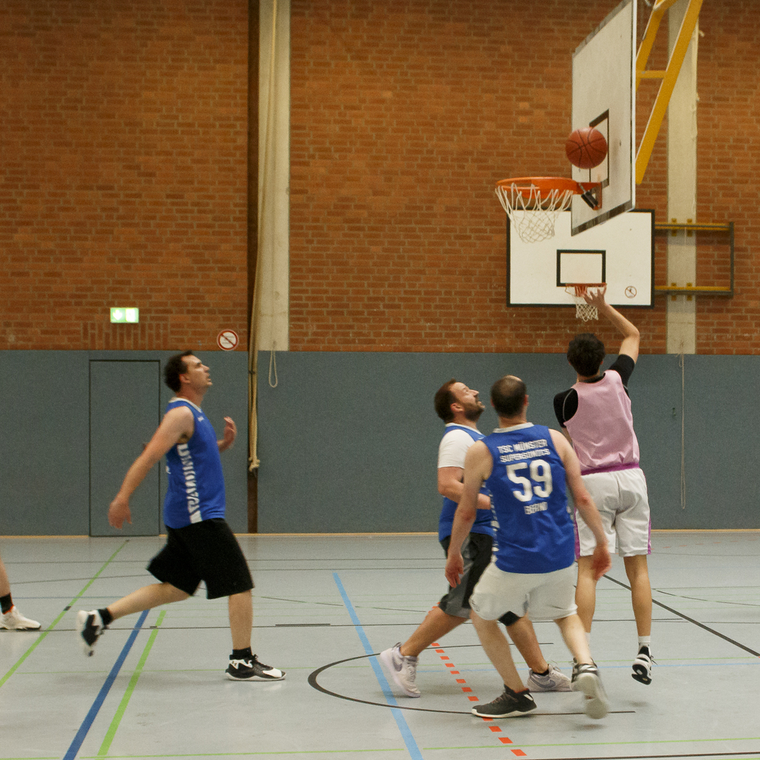 Spielende M&auml;nner in Trikots werfen einen Basketball auf einen Korb in einer Sporthalle.