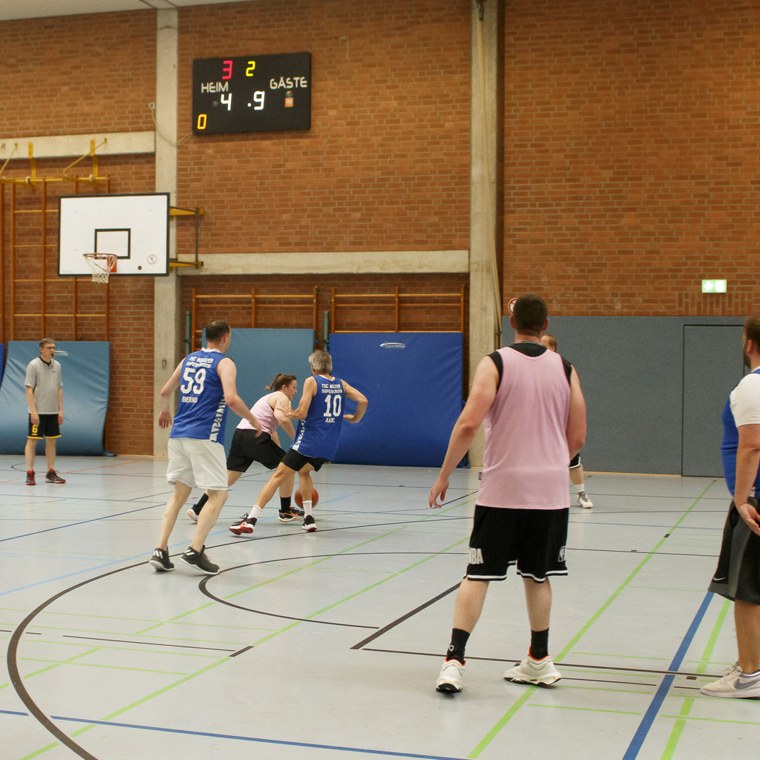 Basketballspiel in der Halle mit Spielern in blauen und pinken Trikots, einem Korb und einer Anzeigetafel im Hintergrund.