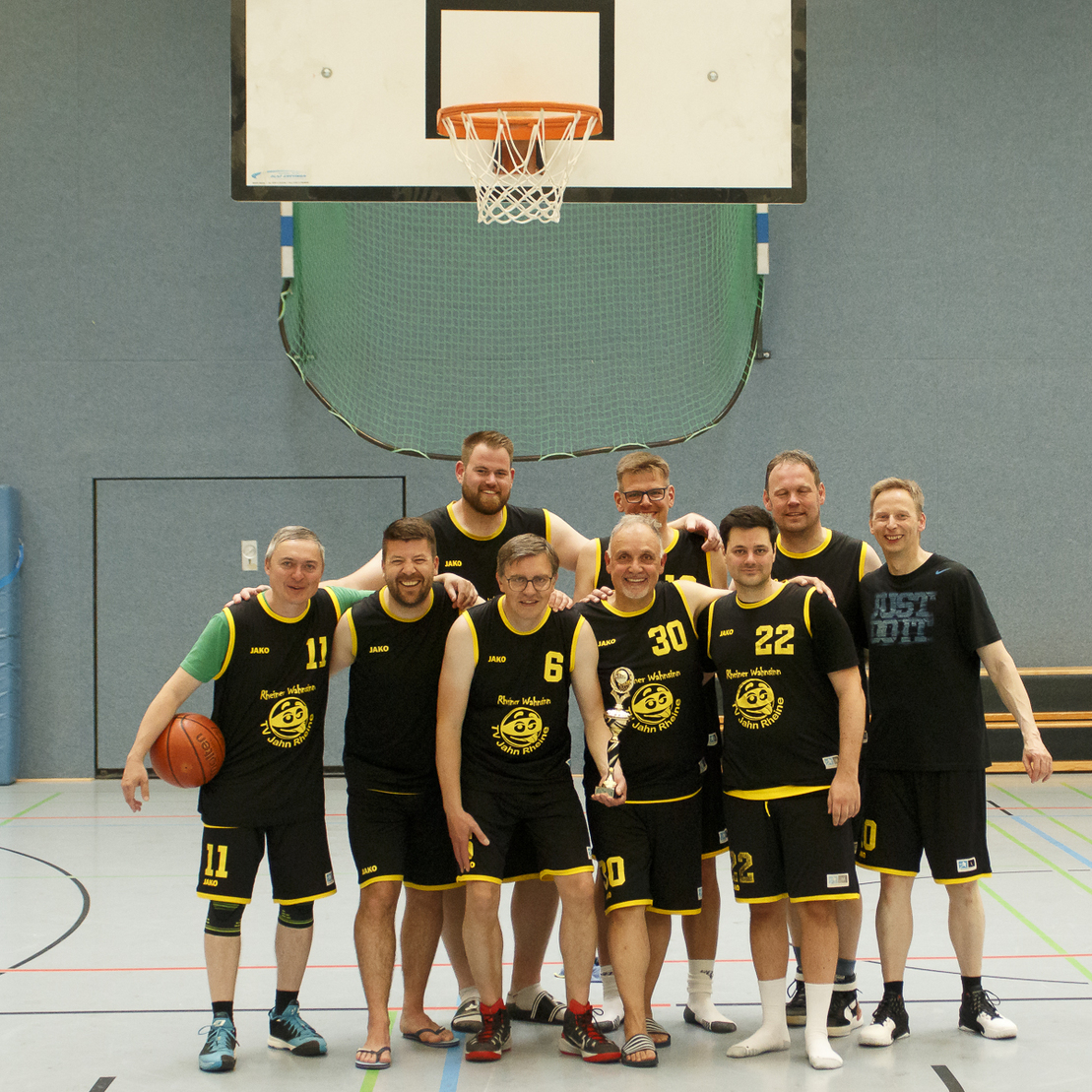 Gruppe von neun m&auml;nnlichen Basketballspielern in schwarzen Trikots, posierend vor einem Korb in einer Sporthalle.