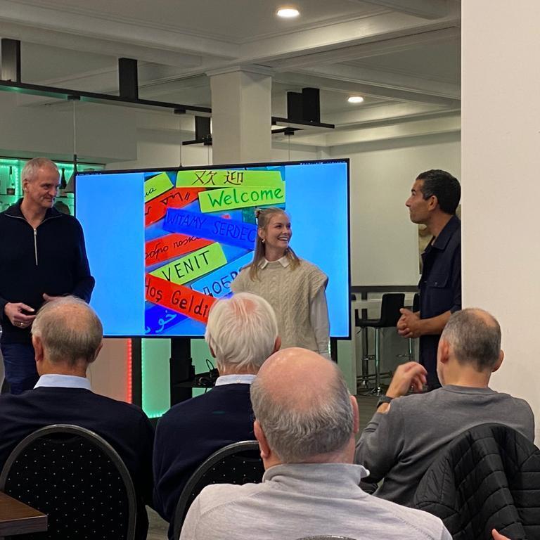 Zwei Personen stehen vor einem Bildschirm mit bunten "Welcome"-Schildern, w&auml;hrend Publikum im Hintergrund zuschaut.