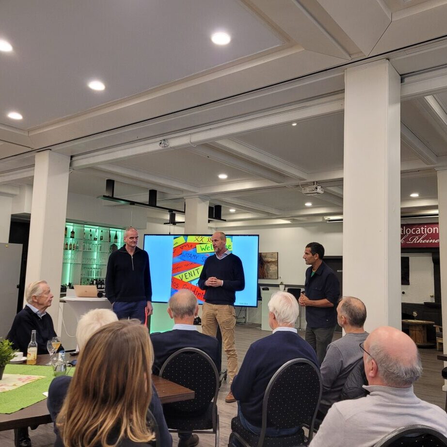 Drei M&auml;nner pr&auml;sentieren vor einem Publikum in einem modernen Raum, Projektionsbildschirm im Hintergrund.