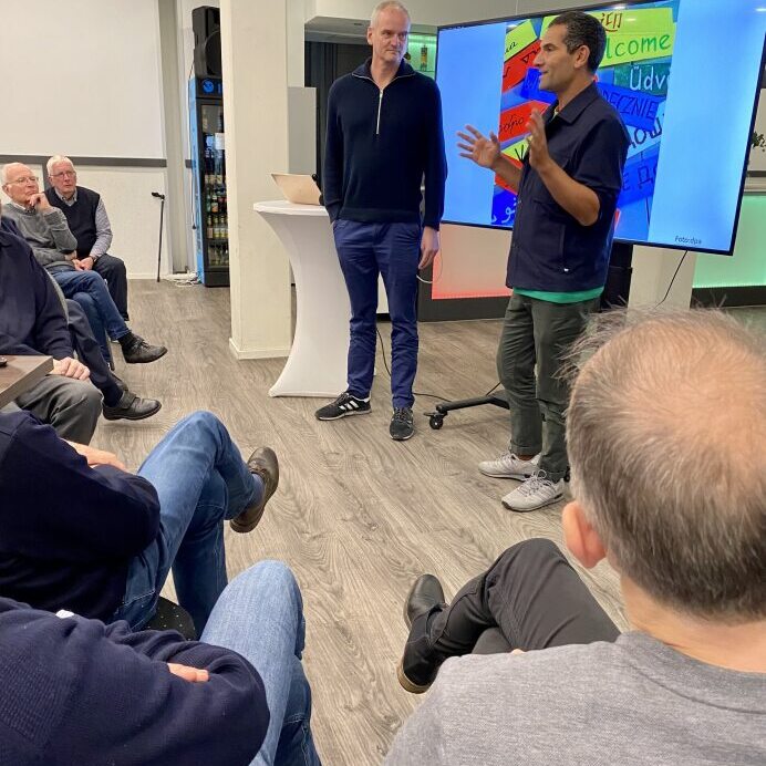 Zwei M&auml;nner sprechen vor einem Publikum, w&auml;hrend ein Bildschirm mit bunten Worten im Hintergrund sichtbar ist.