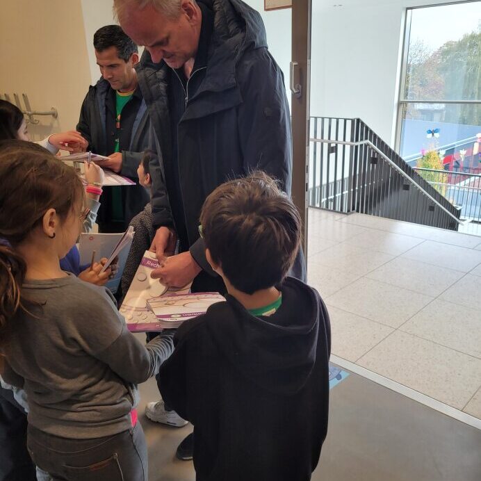 Eine Gruppe von Kindern steht vor einer T&uuml;r und erh&auml;lt von einem Erwachsenen Unterlagen.