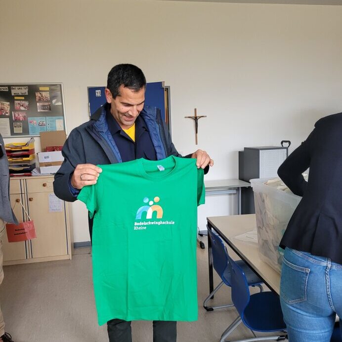 Mann h&auml;lt ein gr&uuml;nes T-Shirt mit dem Logo der Badeschule Rhein in einem Klassenzimmer.