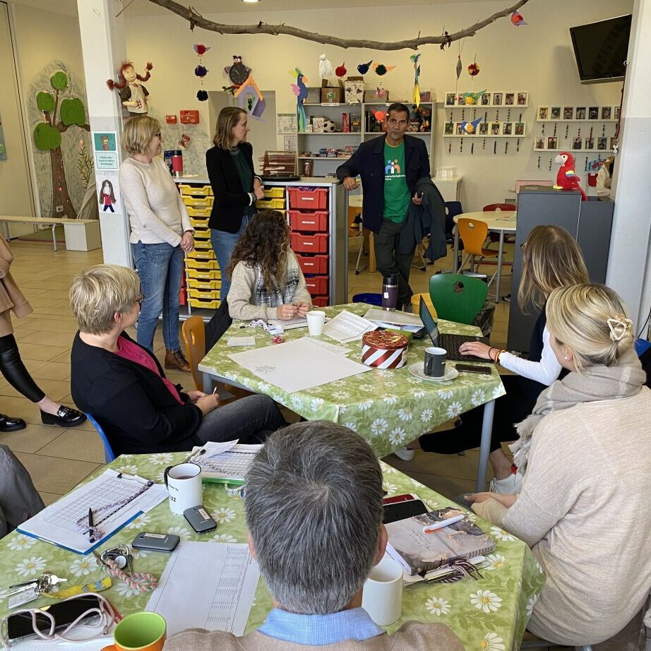 Gruppentreffen in einem Raum mit Teilnehmern um einen Tisch, w&auml;hrend ein Mann steht und spricht.