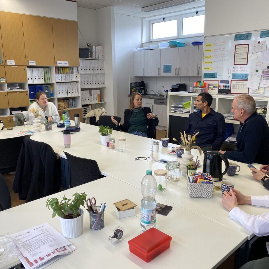 Gruppe von sechs Personen in einem hellen Raum, sitzend um einen Tisch mit Pflanzen und B&uuml;romaterialien.