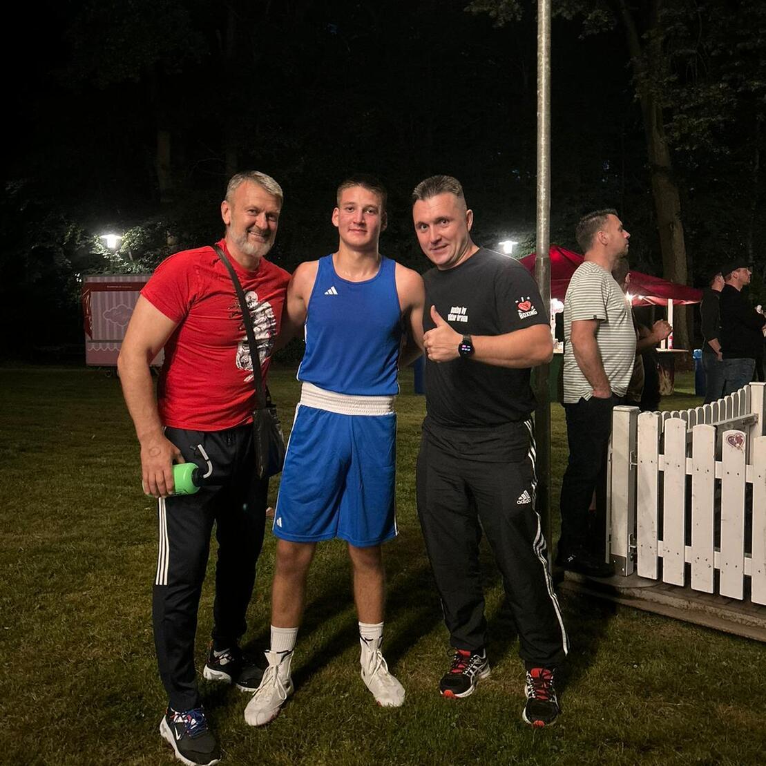 Drei Männer in Sportbekleidung stehen nachts auf einem Rasen, der Boxkämpfer trägt eine blaue Trainingsausrüstung.