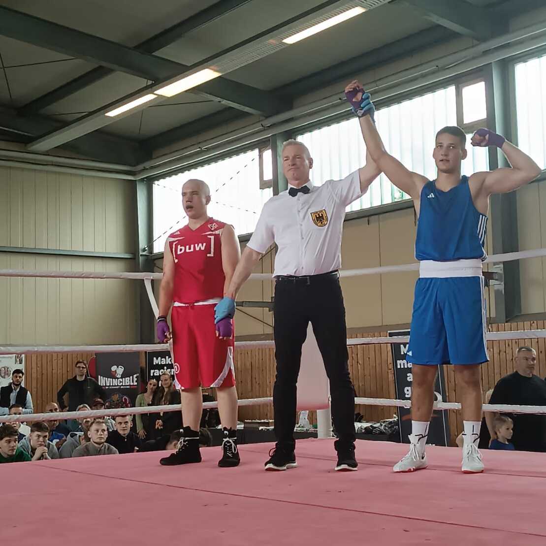 Zwei Boxer in Ringkleidung stehen nebeneinander, während ein Schiedsrichter den Sieger erklärt. Zuschauer im Hintergrund.