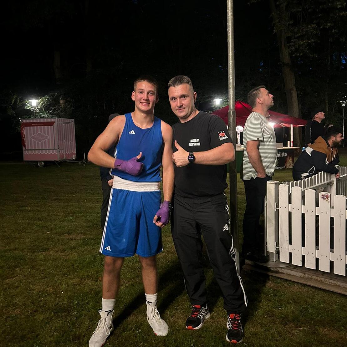 Zwei Männer posieren im Freien bei Nacht; ein Boxer in blauem Outfit mit Handschuhen und ein Trainer mit Daumen hoch.