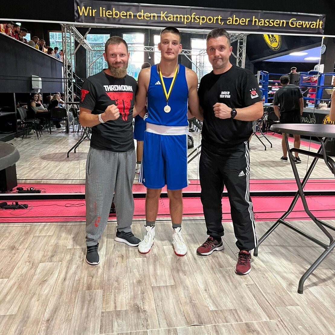 Drei Personen posieren in einem Sportumfeld, der junge Boxer trägt eine Medaille und ein blaues Trikot.