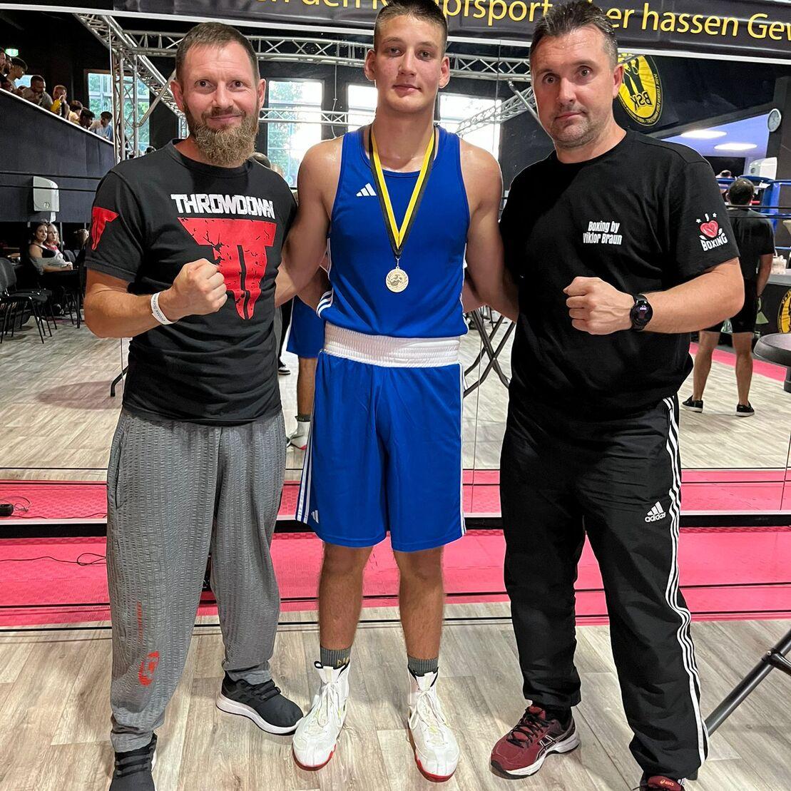 Drei Personen stehen zusammen: Ein Boxer in blauer Sportkleidung mit Medaille, flankiert von zwei Trainern.
