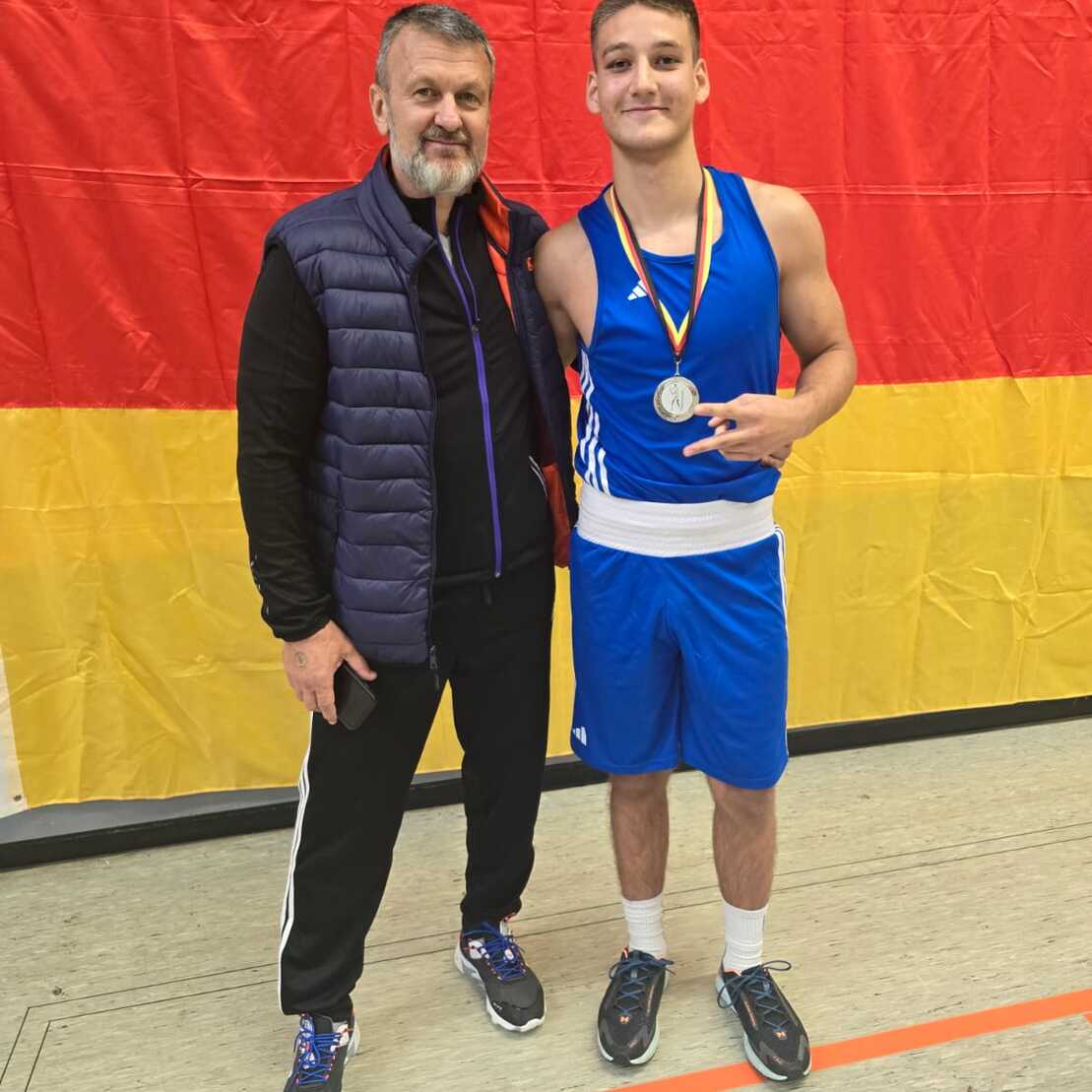 Zwei Personen stehen vor einer deutschen Flagge, einer trägt eine Medaille, beide lächeln in die Kamera.