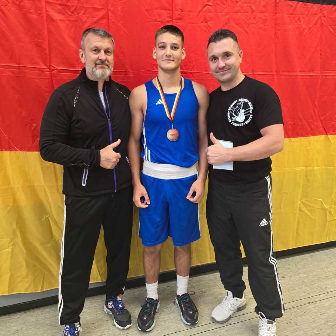 Drei Personen posieren vor einer deutschen Flagge, ein Boxer trägt eine Medaille und steht zwischen zwei Trainern.