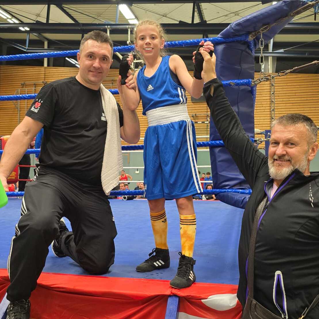 Ein Mädchen im blauen Boxanzug steht in einem Boxring, während zwei Trainer ihre Hände verdienen.