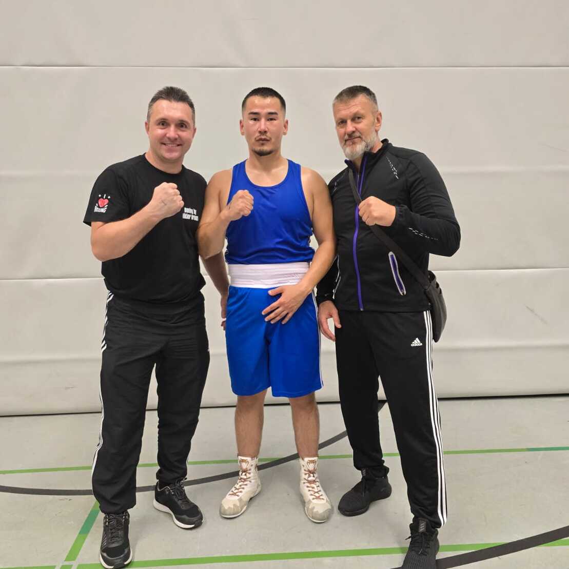 Drei Männer stehen in Sportbekleidung in einer Turnhalle, zwei daneben, einer in der Mitte in Boxausrüstung mit einem Sieger-Lächeln.