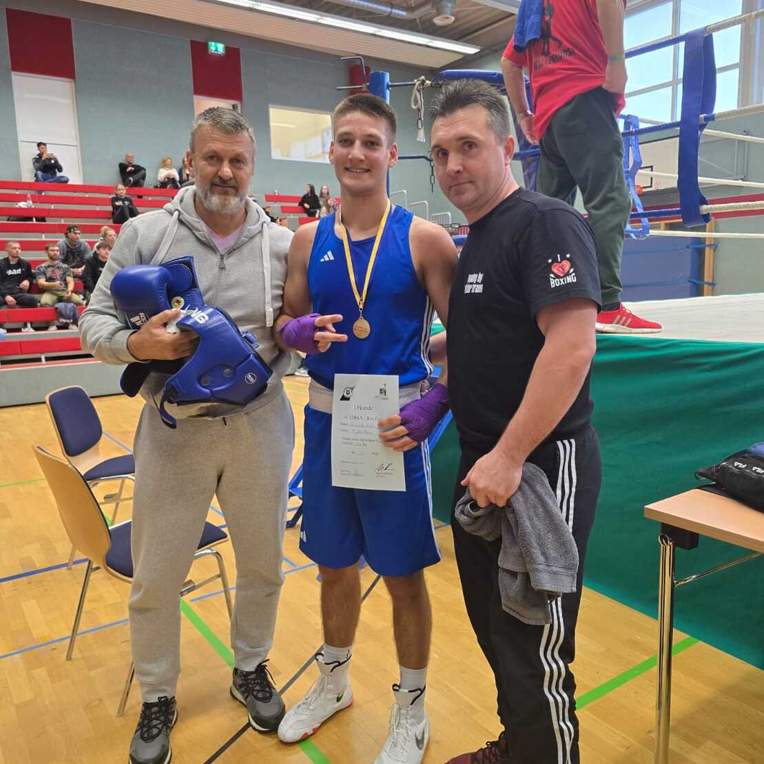 Drei Personen posieren nach einem Boxwettkampf: Ein junger Boxer mit Medaille und Zertifikat, umgeben von zwei Trainern.