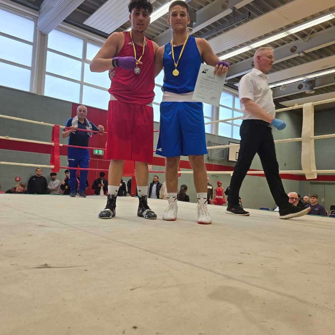 Zwei Boxer in roten und blauen Trikots stehen auf einem Boxring und zeigen stolz ihre Medaillen.