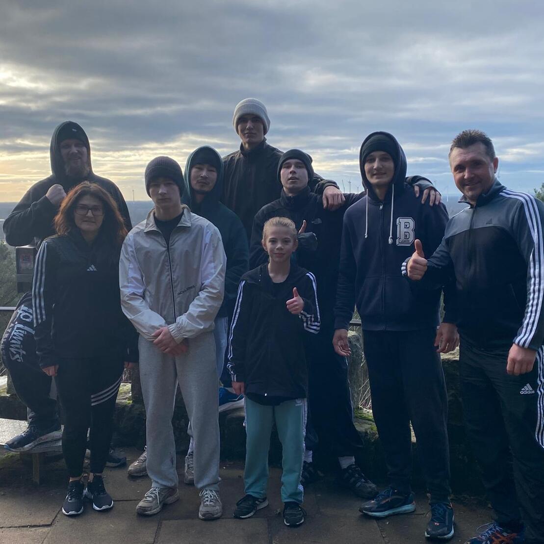 Gruppe von zehn Personen in Sportbekleidung, lächelnd und Daumen hoch, vor einer Aussicht mit Himmel und Bäumen.