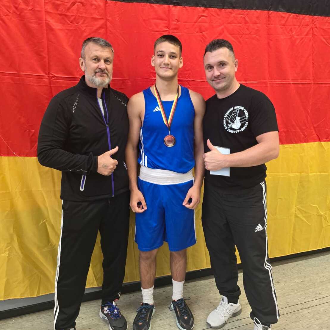 Drei Männer stehen vor einer deutschen Flagge, der jugendliche Boxer trägt eine Bronze-Medaille und lächelt.