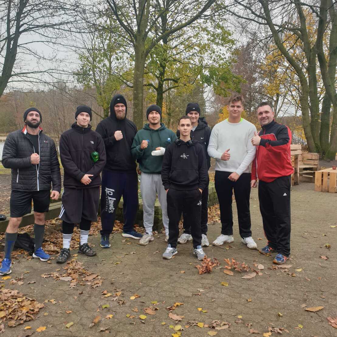 Gruppe von acht jungen Männern in sportlicher Kleidung posiert im Freien umgeben von Bäumen und Laub.
