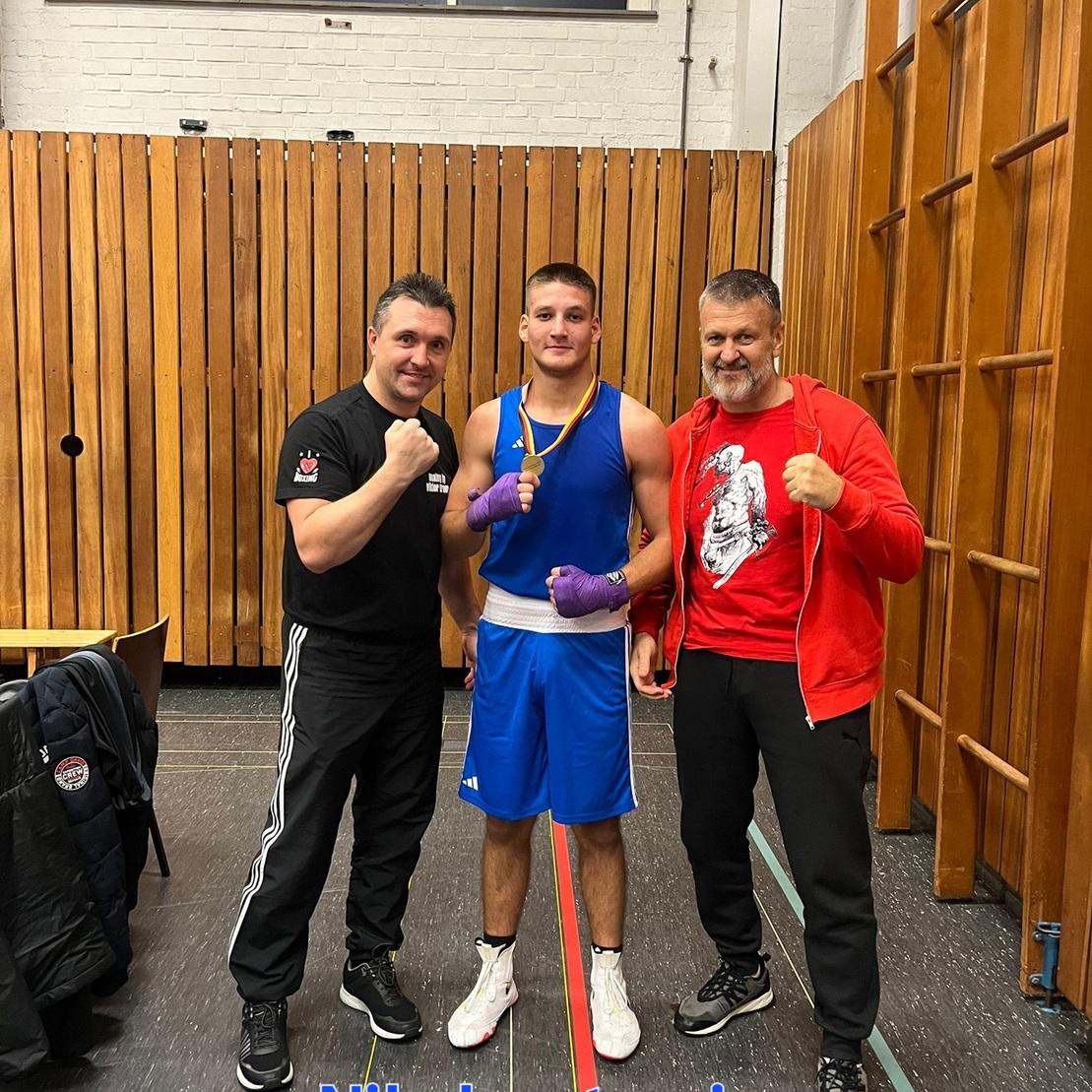 Drei Personen in einem Sportraum nach einem Boxevent, ein Boxer mit Medaille und zwei Trainer in Aktion.