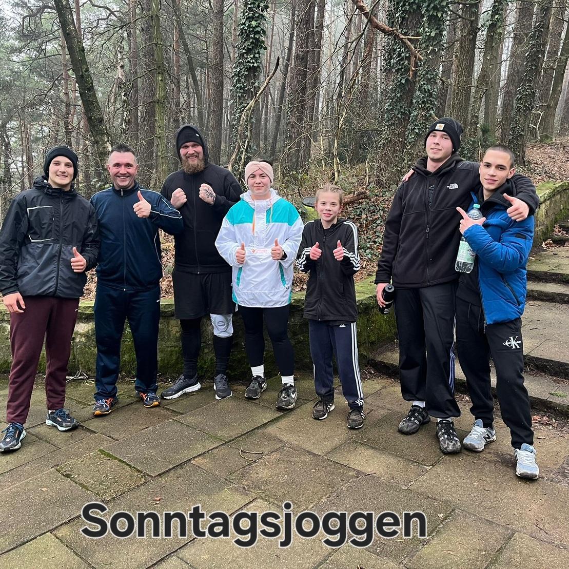 Gruppe von sieben Personen beim Sonntagsjoggen im Wald, alle zeigen Daumen hoch und lächeln.