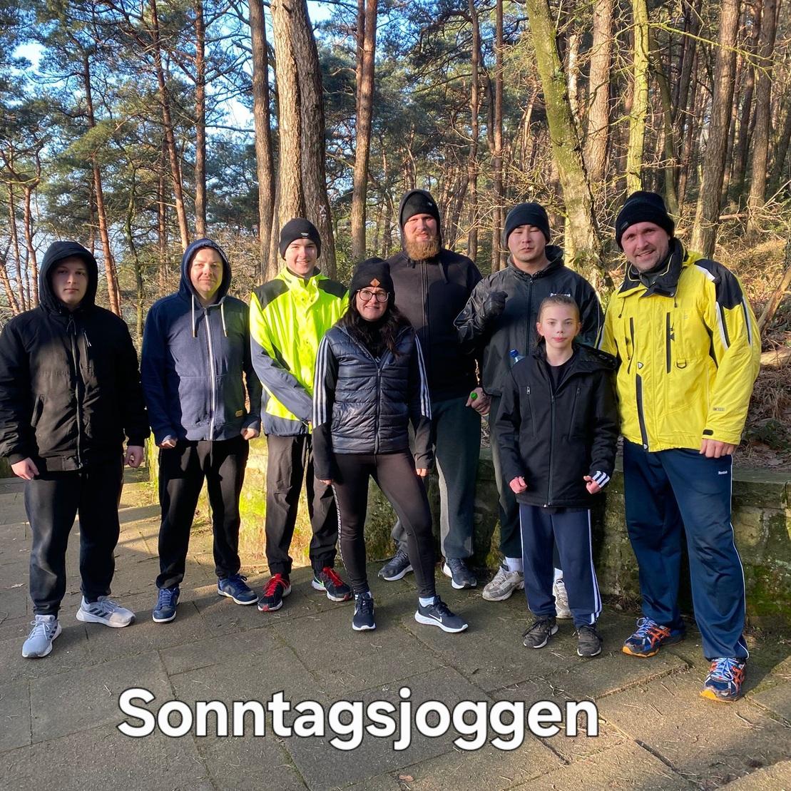 Gruppe von sieben joggenden Menschen in Sportkleidung auf einem Waldweg, umringt von Bäumen. Text: "Sonntagsjoggen".