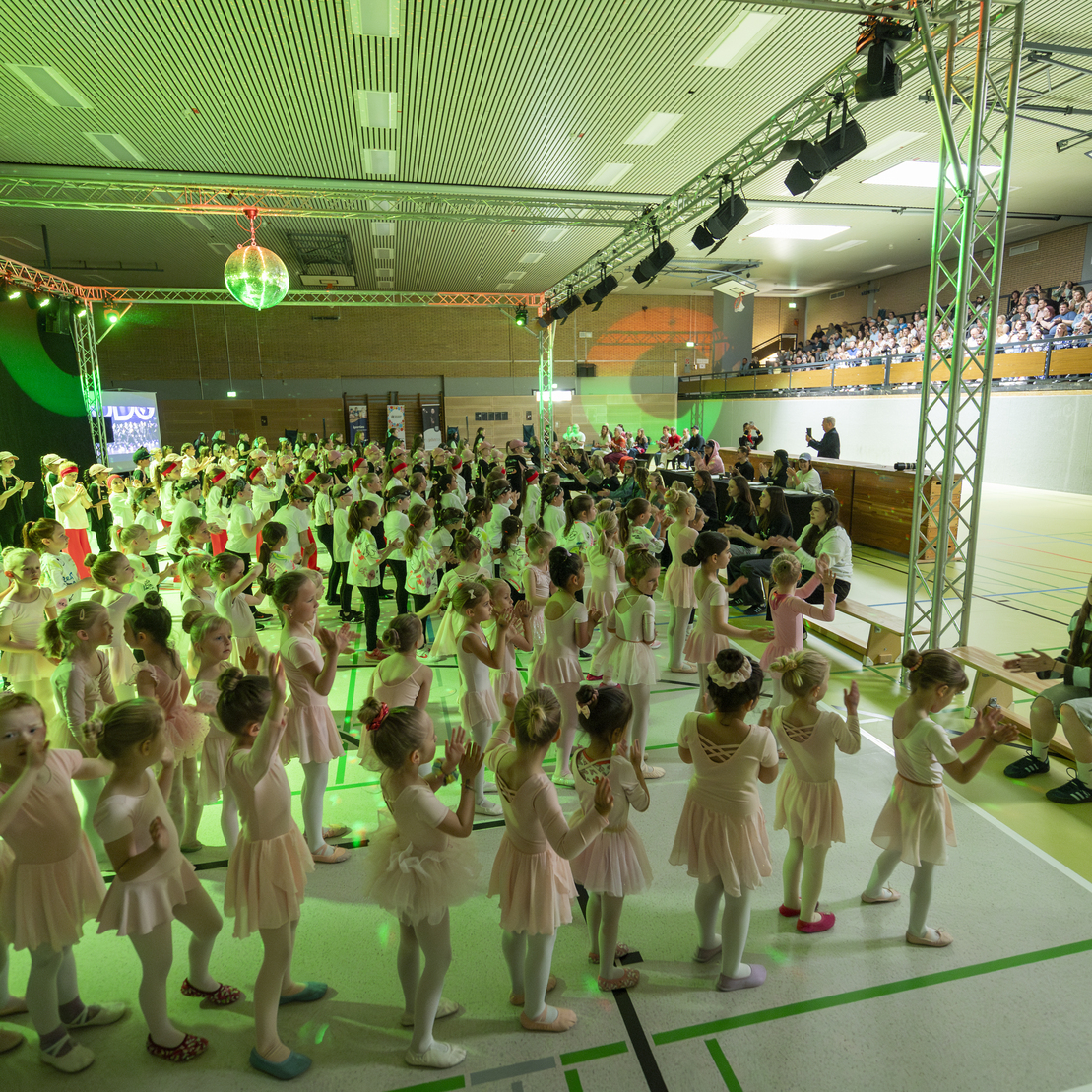 Kindertanzauff&uuml;hrung in einer Halle mit vielen tanzenden Kindern in Ballettkleidung und Publikum im Hintergrund.