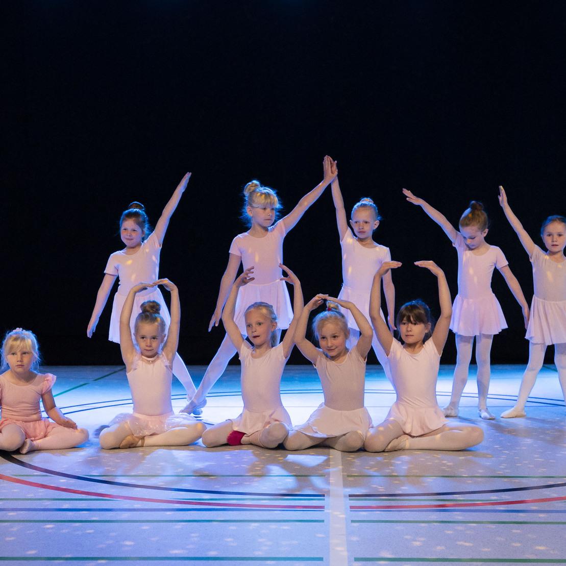 Gruppe von acht Ballettt&auml;nzerinnen in rosa Kost&uuml;men, die in einer choreografischen Pose auf einer Sportfl&auml;che stehen.