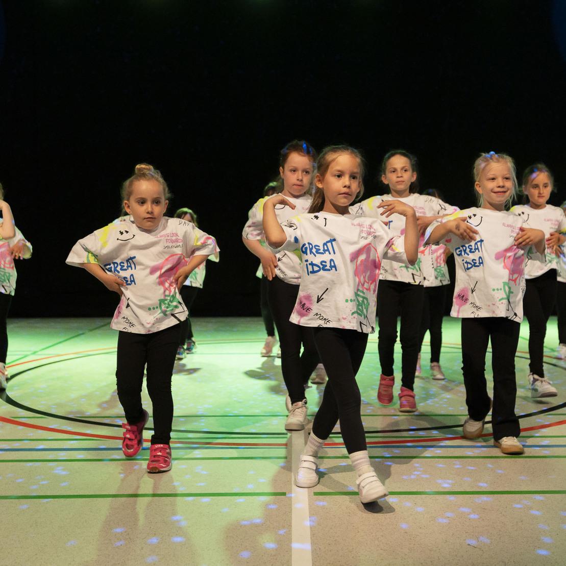 Gruppe von M&auml;dchen in bunten T-Shirts tanzt auf einer beleuchteten Sportfl&auml;che w&auml;hrend einer Auff&uuml;hrung.